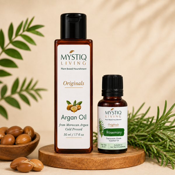 Two bottles of Mystiq Living Originals plant-based nourishment on a wooden stand: a larger amber bottle of Argan Oil and a smaller amber bottle of Rosemary essential oil, styled with argan nuts, green leaves, and rosemary sprigs on a beige background.