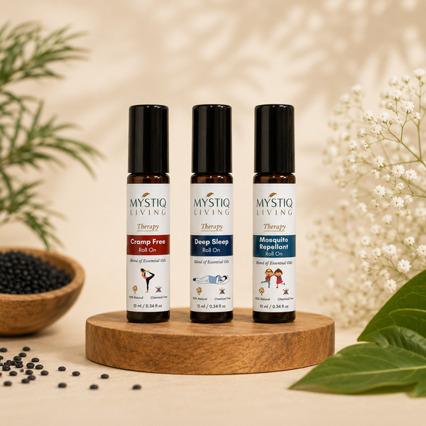 Three small roll-on bottles from Mystiq Living Therapy are displayed on a round wooden stand. The bottles are labeled Cramp Free, Deep Sleep, and Mosquito Repellent, with a neutral beige background decorated with green leaves, white flowers, and a small bowl of black peppercorns.